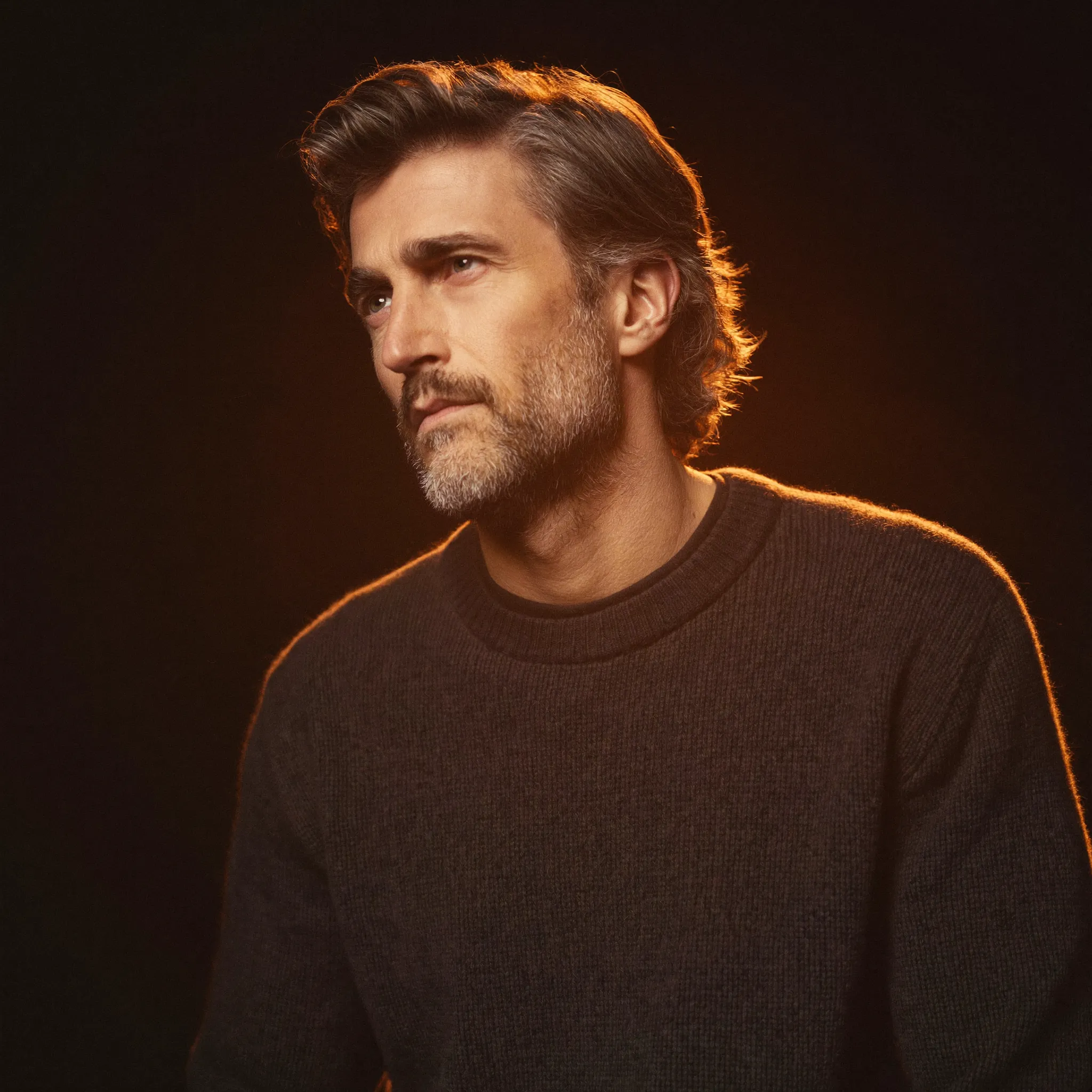 Portrait of Johan, a White agency owner with a salt-pepper beard who sells design retainers, wearing a charcoal FantoClient sweatshirt in warm candlelight